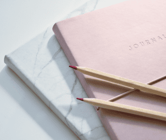 Two closed notebooks, one pink and one with a marble pattern, are stacked elegantly on a surface, showcasing sophisticated branding. Two sharpened red-tipped pencils rest on top, complementing the stylish ensemble.