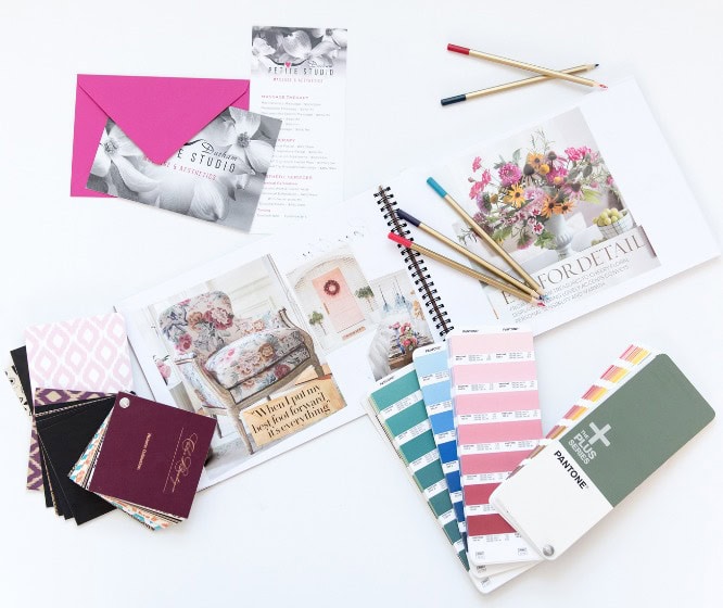 An open interior design book with floral images, fabric swatches, paint color samples, and pencils sits on a white surface, alongside a bright pink envelope showcasing the essence of creative branding.