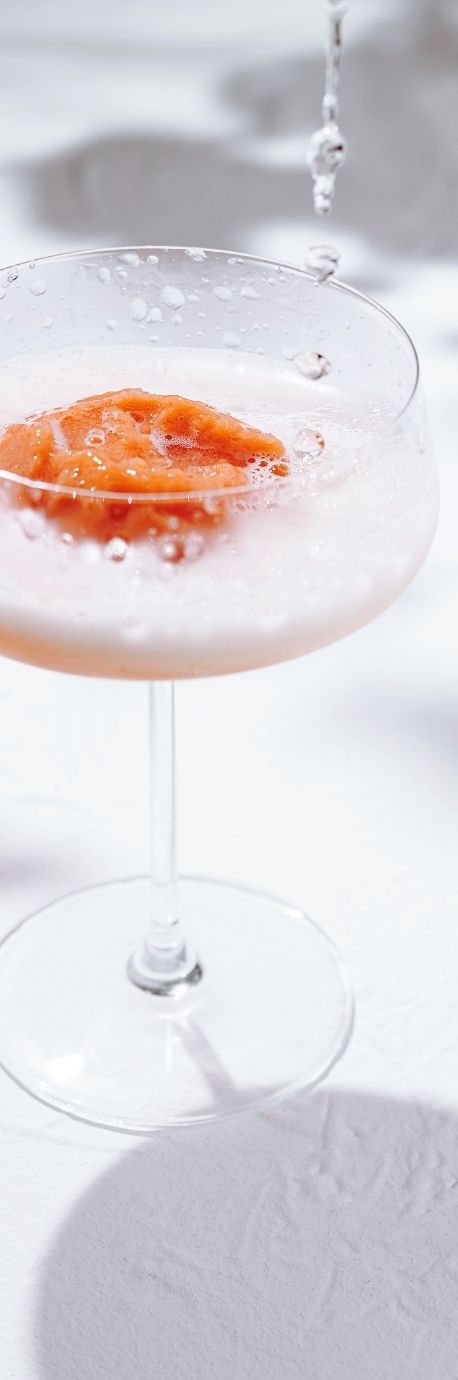 A frothy cocktail in a glass with an orange slice garnish, artfully arranged on a white surface with shadows, reflecting Sue Forrest's distinctive brand design.