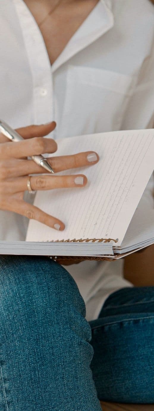 A person in a white shirt and jeans writes in a spiral-bound notebook with a silver pen, capturing thoughts on brand evolution.