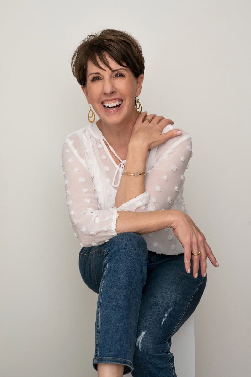 Meet Sue Forrest: a woman wearing a white blouse and jeans, smiling while seated against a plain background.