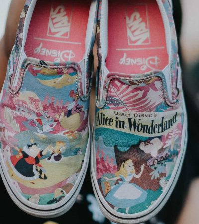 A person holds up a pair of slip-on shoes with a colorful "Alice in Wonderland" design featuring various cartoon characters.