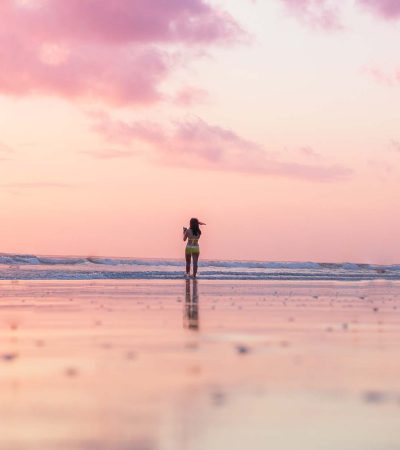 A person stands on a wide beach at sunset, embraced by pink clouds and gentle waves, feeling the self-empowerment that comes with knowing they are enough.