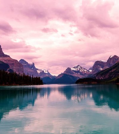 In 2022, a tranquil lake, reflecting snow-capped mountains under a pink-tinged cloudy sky, is surrounded by dense forests and rugged peaks.